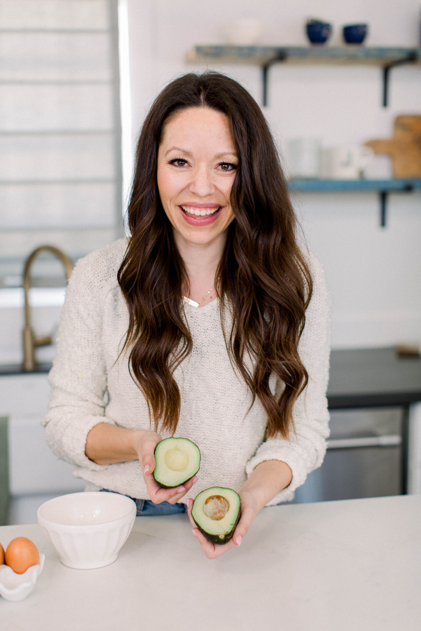 a smiling Chelsi jo holds a cut avocado in a crisp white kitchen