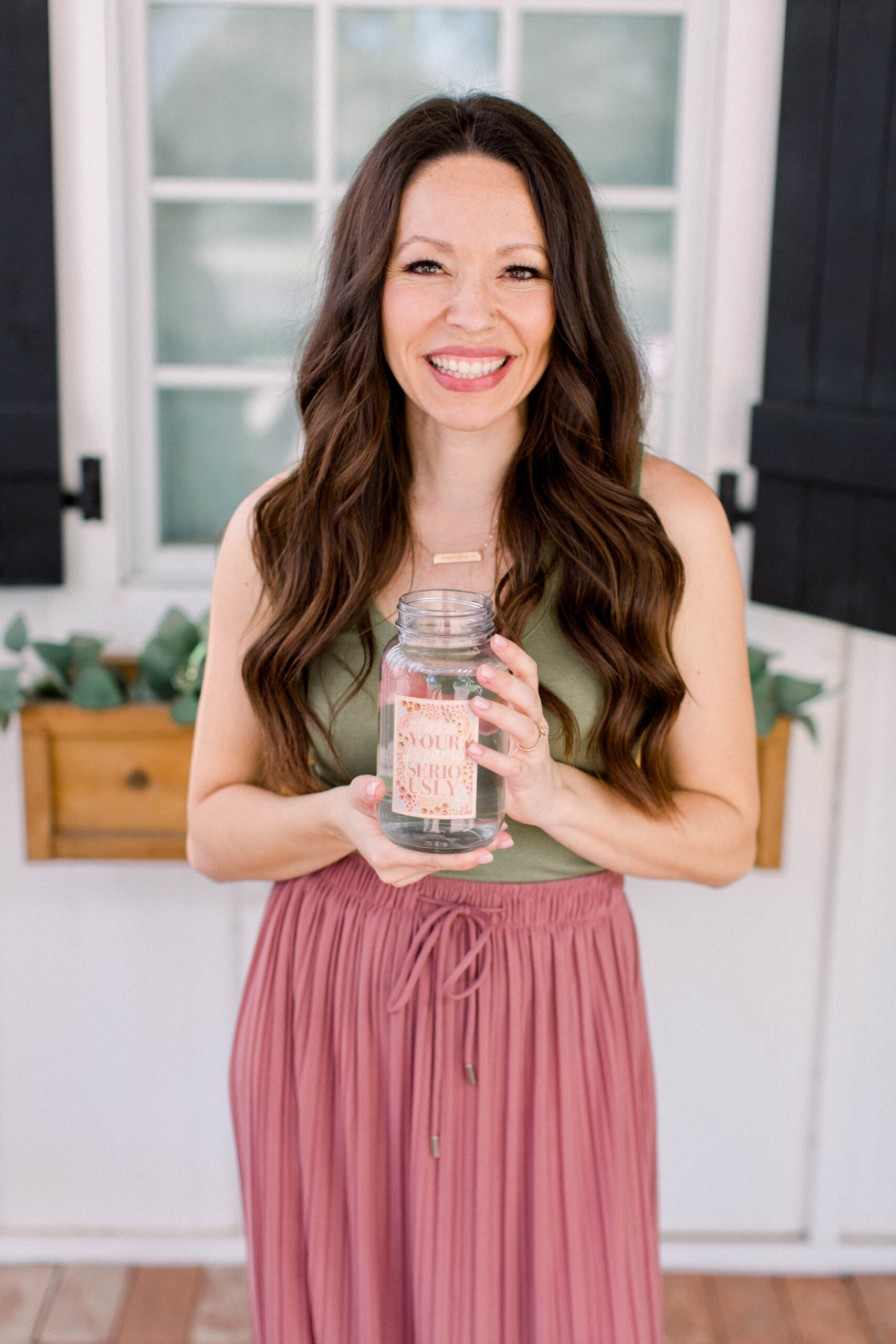 Chelsi Jo smiles into the camera holding a mason jar, wearing an olive green tank and a dusty pink skirt